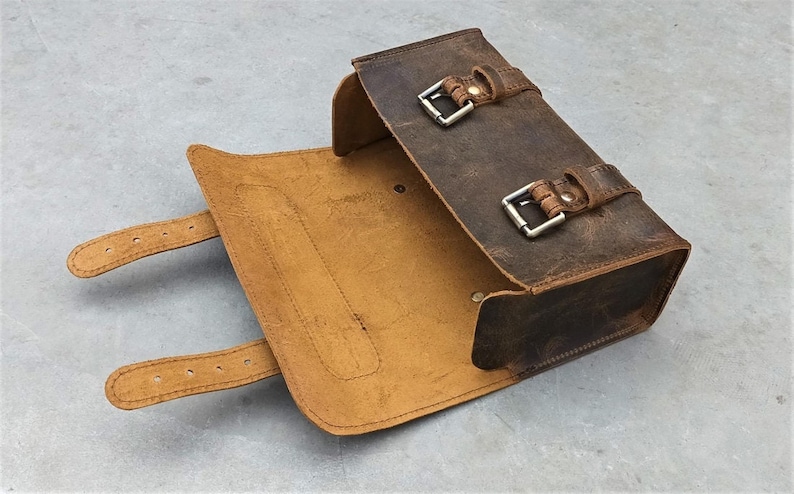 A brown leather tool bag is shown open with its flap down and buckle straps extended on a gray background.