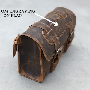 A brown leather motorcycle tool bag angled on a gray surface with an arrow pointing to the flap for custom engraving.