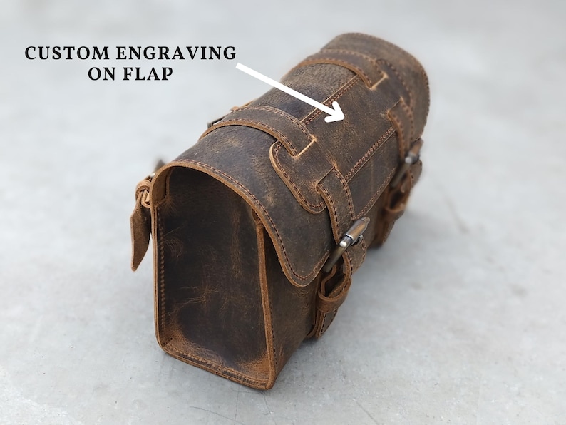 A brown leather motorcycle tool bag angled on a gray surface with an arrow pointing to the flap for custom engraving.