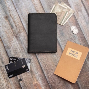 Puede incluir: Un cuaderno de cuero negro con un borde cosido marrón, un cuaderno marrón con el texto "FIELD NOTES" en la portada, dos tarjetas de crédito y algunos billetes sobre una superficie de madera.