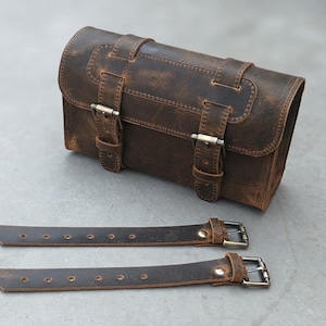 A brown leather motorcycle tool bag is displayed next to two separate leather straps with metal buckles.
