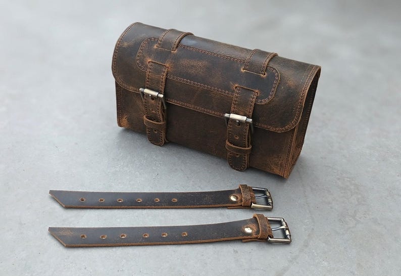 A brown leather motorcycle tool bag is displayed next to two separate leather straps with metal buckles.