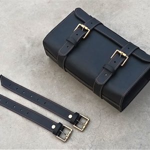 Può includere: Borsa in pelle nera con due cinturini regolabili e fibbie dorate. La borsa ha una forma rettangolare ed è realizzata in pelle di alta qualità.