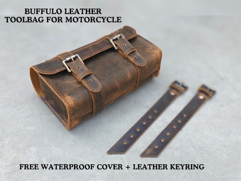 A brown leather motorcycle tool bag lies on a gray surface with two matching mounting straps placed beside it.