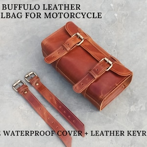 Pode incluir: Uma bolsa de ferramentas de couro castanho para motocicleta com duas correias e fivelas de metal. O texto no topo diz "BUFFULO LEATHER TOOLBAG FOR MOTORCYCLE". O texto na parte inferior diz "FREE WATERPROOF COVER + LEATHER KEYRING".