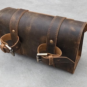 A brown leather motorcycle tool bag is shown resting on a flat surface with its buckle straps wrapped around the body.