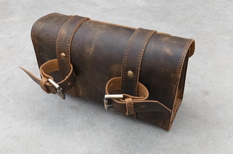 A brown leather motorcycle tool bag is shown resting on a flat surface with its buckle straps wrapped around the body.