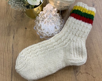 Puede incluir: Calcetín tejido a mano de color crema, con un patrón de canalé y un puño con rayas amarillas, verdes y rojas. Una madeja de lana y agujas de tejer están junto al calcetín. Un pequeño arreglo floral y un árbol decorativo de cristal también están en la imagen.