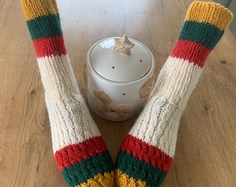 Puede incluir: Un par de calcetines tejidos a mano con un diseño de bloques de color. Los calcetines presentan bandas amarillas, verdes y rojas en el puño y la puntera, con un cuerpo color crema. Un tarro de cerámica blanco con tapa en forma de estrella y diseño de caballito balancín está en el fondo.