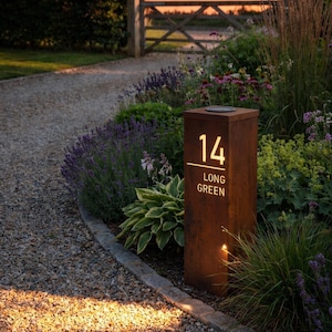 May include: A rectangular, weathered rust-effect metal house number sign in a vertical post style. The sign is internally illuminated, showing the number 14 above the text LONG GREEN in a clear, sans-serif typeface, situated within a garden.