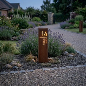 May include: A rectangular, rust-coloured metal outdoor house number sign featuring the number 14 and the text LONG GREEN illuminated in warm light. The vertical post stands in a garden bed with lavender plants and gravel, serving as a marker.