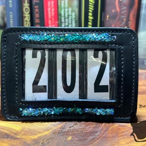 May include: Black leather accessory with the numbers "202" displayed in a clear window. The frame is embellished with turquoise and teal colored rhinestones. The accessory is likely a calendar or display piece.