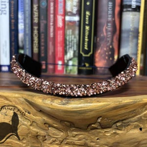 May include: A black headband adorned with rose gold crystals. The crystals are arranged across the top of the headband in a curved pattern. The headband is displayed on a wooden surface, with a bookshelf in the background.