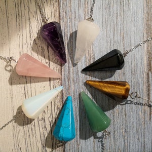 Puede incluir: Un conjunto de nueve péndulos de cristal, cada uno con un color y tipo de cristal diferente. Los péndulos cuelgan de cadenas plateadas y están dispuestos en círculo sobre una superficie de madera.