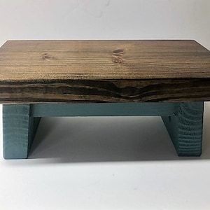 May include: A small wooden stool with a dark brown top and teal blue legs. The stool is made of solid wood and has a rustic, farmhouse style.