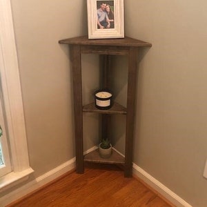May include: A wooden corner shelf with three tiers. The top tier has a white picture frame with a photo of a couple. The middle tier has a black candle. The bottom tier has a small potted plant.