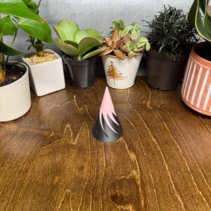 May include: A two-toned, pink and black cone-shaped object sits on a wooden surface. The object has a pointed top and a flared base with cut-out designs. Various potted plants are in the background.