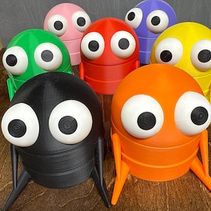 May include: A collection of colorful, 3D-printed creature-shaped containers. Each container features large, white-rimmed eyes with black pupils and small, leg-like appendages. The containers are in various colors, including green, pink, red, purple, yellow, orange, and black.