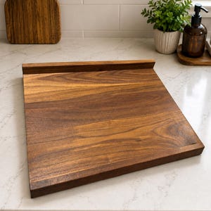 May include: A square wooden cutting board made of dark brown walnut wood with a visible natural grain pattern. The board features a raised edge along the back side, providing a ledge for stability or support while in use on a flat surface.