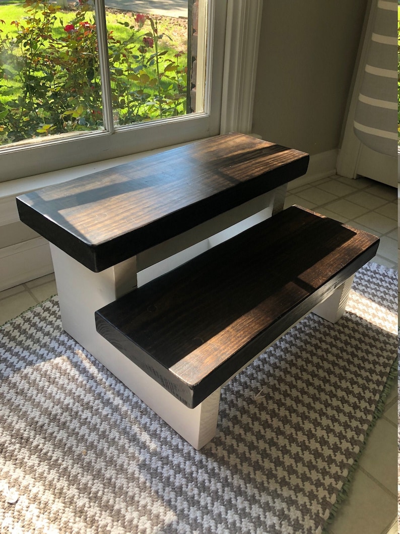 May include: A two-step wooden stool with a white base and dark brown stained top steps. The stool is sitting on a white and gray patterned rug.