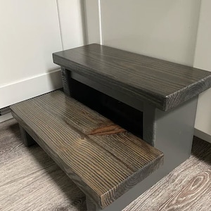 May include: A two-step wooden stool with a dark gray stain. The top step is wider than the bottom step. The stool is made of solid wood and has a rustic look.