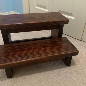 May include: A dark brown wooden step stool with two steps. The top step is wider than the bottom step.