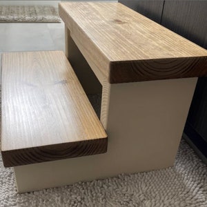 May include: A two-step wooden stool with a natural wood top and a white painted base. The stool has a simple design and is perfect for reaching high shelves or adding extra seating.