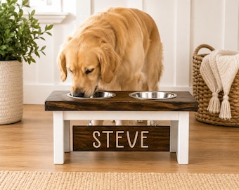 May include: A raised wooden pet feeder with a dark brown top and white legs, featuring two stainless steel bowls. A rectangular wooden sign hangs from the center of the frame with the name STEVE engraved in light-colored capital letters.