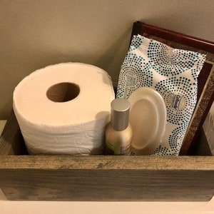 May include: A rustic wooden box with a roll of toilet paper, a package of facial tissues, a small bottle of hand sanitizer, and a disposable plate. The box is gray and has a distressed finish.