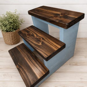 May include: A three-step wooden stool featuring dark brown stained treads with visible wood grain and light blue painted side supports. The structure has a tiered design with three distinct levels, providing a functional step arrangement.