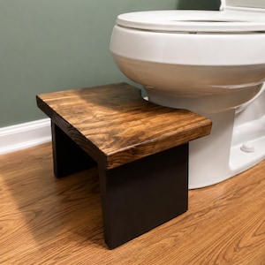 May include: A rectangular wooden step stool featuring a dark brown stained top surface with visible wood grain and solid black painted side supports. The stool has a curved cutout along one side to fit against the base of a white toilet.