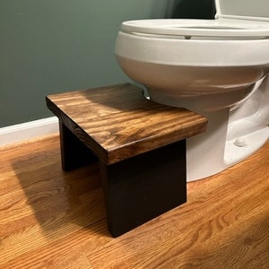 May include: A wooden step stool with a dark brown top and black painted legs. The stool is in a bathroom next to a white toilet.