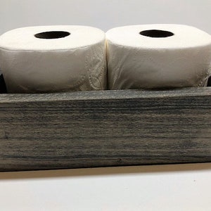 May include: A rustic wooden box with a gray wash finish, designed to hold two rolls of toilet paper. The box has a rectangular shape and a simple design.