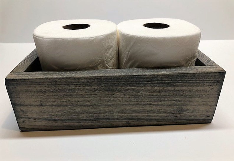May include: A rustic wooden box with a gray wash finish, designed to hold two rolls of toilet paper. The box has a rectangular shape and a simple design.