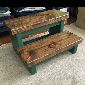 May include: A two-step wooden stool with a dark brown stain and a forest green painted base. The stool has a rectangular top and a smaller rectangular step below.