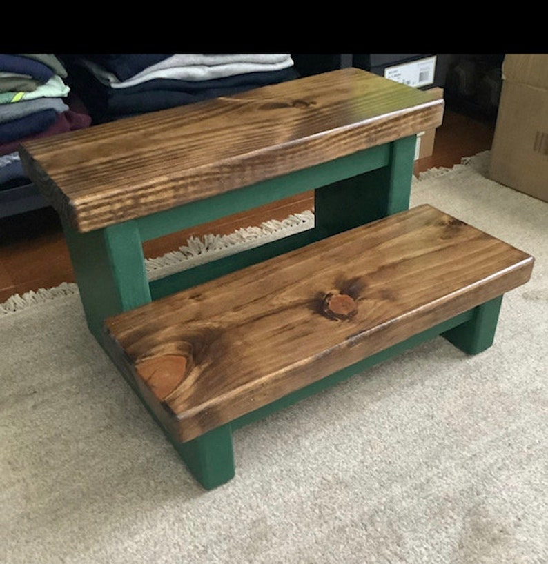May include: A two-step wooden stool with a dark brown stain and a forest green painted base. The stool has a rectangular top and a smaller rectangular step below.