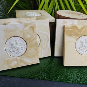 May include: A collection of square coasters with a cream and tan marbled design. Each coaster features a white circular emblem with a detailed relief of a figure on a horse. The coasters are arranged on a dark green surface.