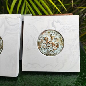 May include: Square marble coasters with a bronze-colored medallion featuring a detailed relief of a figure on a horse. The coasters have a white and gray marbled pattern.