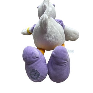 May include: A plush Daisy Duck toy, viewed from the back, with white feathers, purple feet, and a purple top. The toy has yellow accents on the feet and wrists. The Disney licensed toy has a circular logo on the bottom of the left foot.