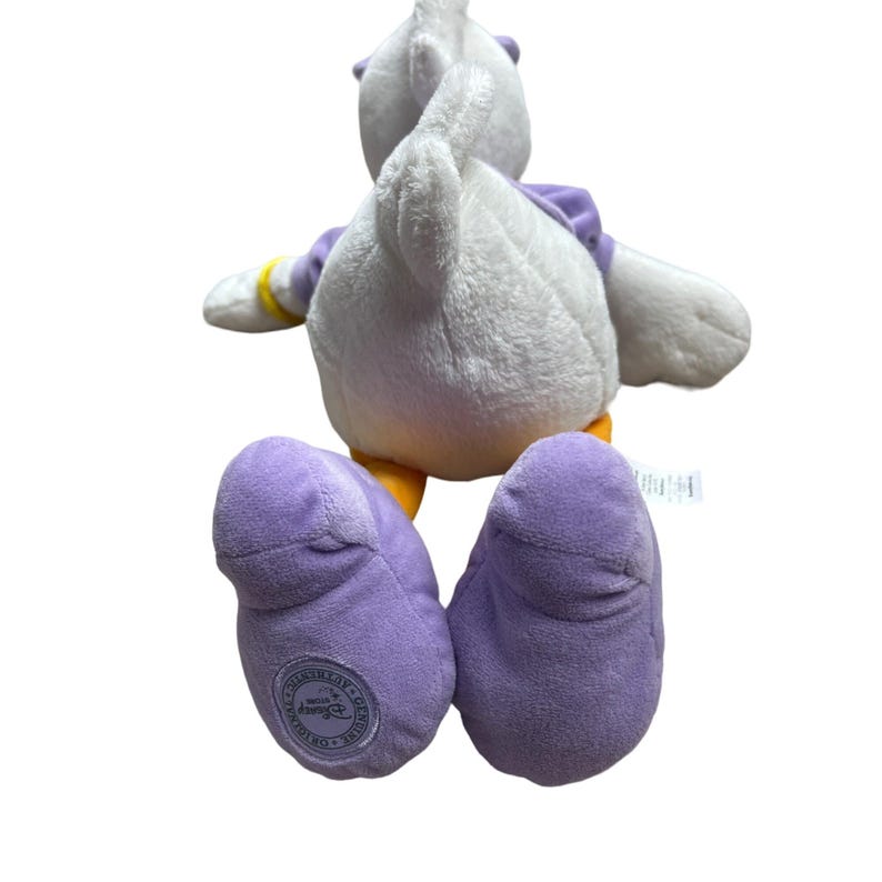 May include: A plush Daisy Duck toy, viewed from the back, with white feathers, purple feet, and a purple top. The toy has yellow accents on the feet and wrists. The Disney licensed toy has a circular logo on the bottom of the left foot.