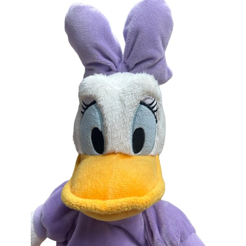 May include: A plush Daisy Duck doll with a white head, blue eyes, and a yellow beak. The doll has purple ears and a purple dress. The doll is made of soft, plush material.