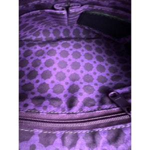 May include: Purple fabric with a geometric pattern lining the inside of a bag. The fabric has a zipper with a silver pull.