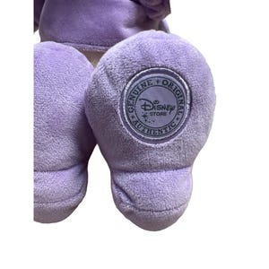 May include: Close-up of a lavender plush toy's foot with a circular Disney Store Authentic seal. The plush is made of soft, fuzzy material. The seal features the Disney logo and the words "Genuine Original" around the edge.