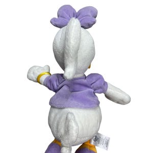 May include: A plush Daisy Duck doll, viewed from the back, features a white body, a purple bow, and a matching purple top. Gold accents are visible on the wrist and feet. The doll has a tag attached to the bottom.