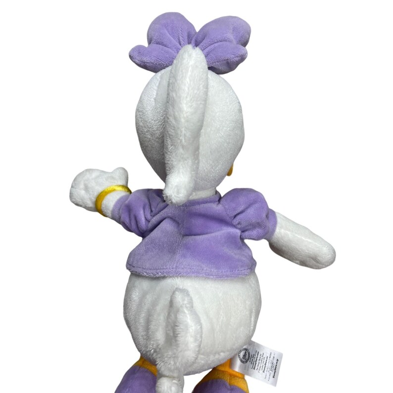 May include: A plush Daisy Duck doll, viewed from the back, features a white body, a purple bow, and a matching purple top. Gold accents are visible on the wrist and feet. The doll has a tag attached to the bottom.