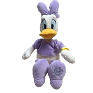 May include: A plush Daisy Duck doll with a white face, a yellow beak, and large blue eyes. She wears a purple top and matching shoes. Her arms and feet are white. The doll has a purple bow on her head and a gold bracelet.