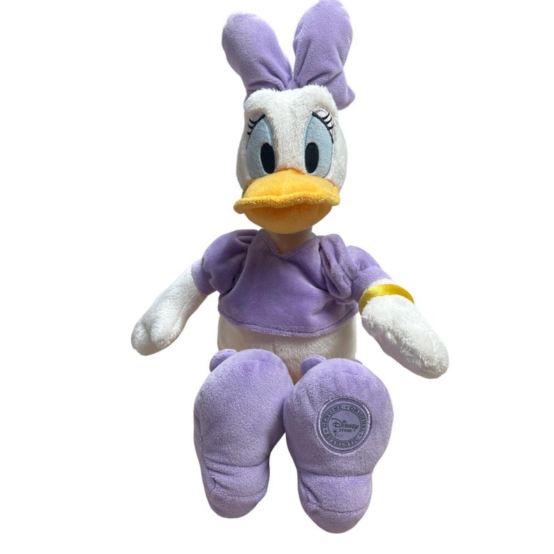 May include: A plush Daisy Duck doll with a white face, a yellow beak, and large blue eyes. She wears a purple top and matching shoes. Her arms and feet are white. The doll has a purple bow on her head and a gold bracelet.