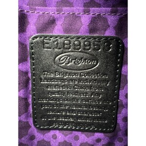 May include: A black leather tag with white stitching and the text "E18995" and "Brighton" in a white circle. The text "The Brighton Collection Handbags are individually crafted in China from quality leather.  All Brighton products are guaranteed against defects in materials and workmanship.  See the Brighton Guarantee for full details." is printed in white.