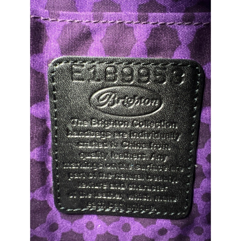 May include: A black leather tag with white stitching and the text "E18995" and "Brighton" in a white circle. The text "The Brighton Collection Handbags are individually crafted in China from quality leather.  All Brighton products are guaranteed against defects in materials and workmanship.  See the Brighton Guarantee for full details." is printed in white.