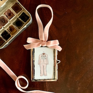 May include: A small, rectangular, silver-toned frame with a pink velvet ribbon and bow. Inside the frame is a watercolor illustration of pink striped pajamas hanging on a hanger, with the words "Do Not Disturb".